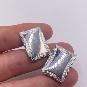 May include: A pair of rectangular silver cufflinks. Each cufflink has a slightly curved, reflective center and a detailed, engraved border. The design features a symmetrical pattern of curved lines and small dots.