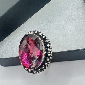 May include: A large, oval-shaped ring with a faceted, magenta-colored gemstone. The gemstone is set in a silver-toned metal band with a beaded border. The ring is displayed inside a black jewelry box with a white interior.