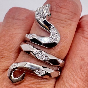 May include: A silver snake ring with a coiled design. The ring features a snake head with a small green stone and is encrusted with small, sparkling white stones. The ring wraps around the finger in a spiral pattern.