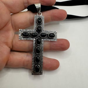 Op de afbeelding: Een zilveren kruis hanger met zwarte ovale stenen die in het midden van elke arm zijn gezet. Het kruis is op een zwart lint.