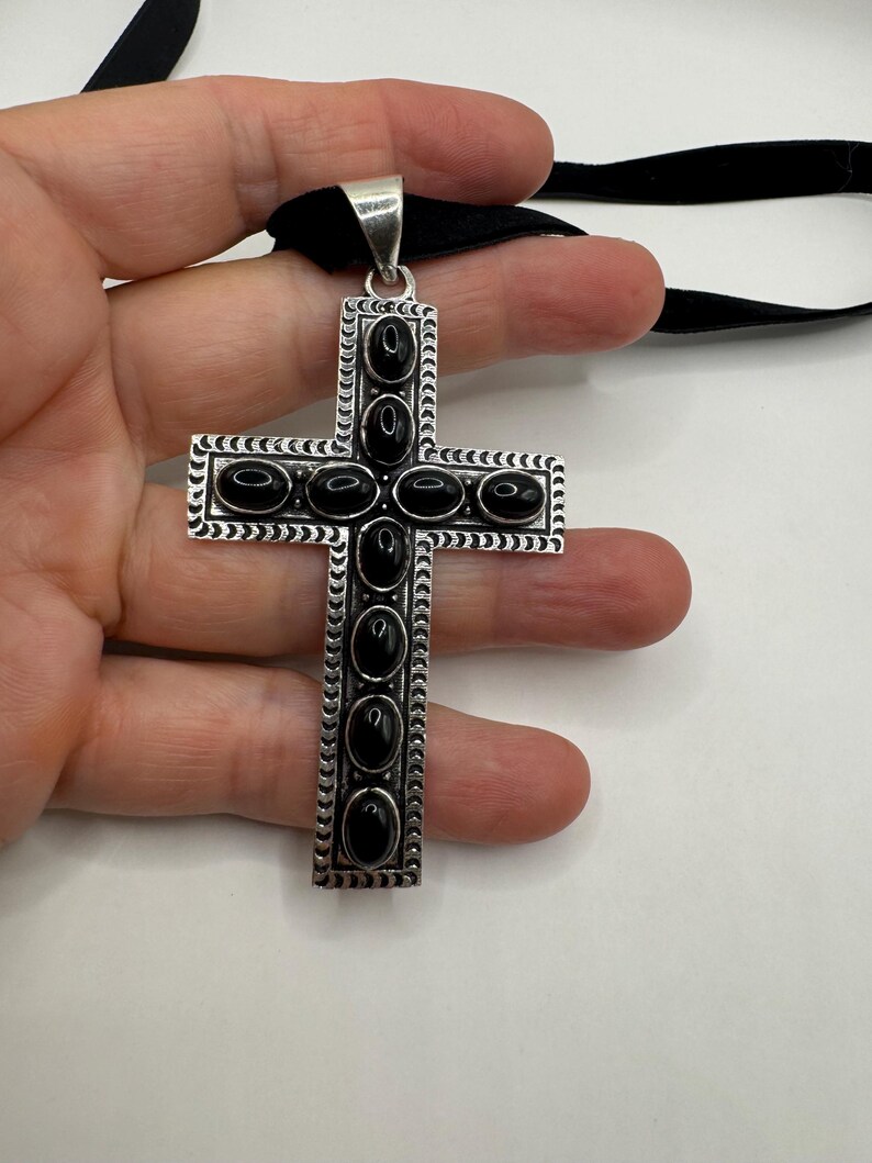 Op de afbeelding: Een zilveren kruis hanger met zwarte ovale stenen die in het midden van elke arm zijn gezet. Het kruis is op een zwart lint.