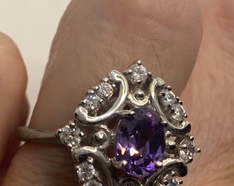 May include: A silver ring with a purple gemstone surrounded by smaller clear gemstones. The ring has a delicate, ornate design.