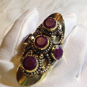 Puede incluir: Un anillo de oro con cinco piedras preciosas púrpuras engastadas en un diseño decorativo. El anillo está diseñado para llevarse en el nudillo.