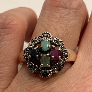 Puede incluir: Un anillo de plata con un grupo de piedras preciosas de colores engastadas en un diseño de marcasita. El anillo presenta piedras preciosas verdes, rojas y negras.