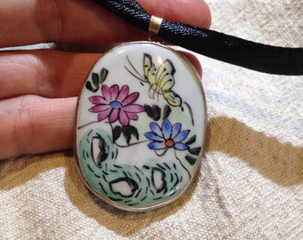 Può includere: Pendente ovale in porcellana con un nastro nero. Il pendente presenta un design dipinto a mano di una farfalla e tre fiori in tonalità rosa, blu e giallo.