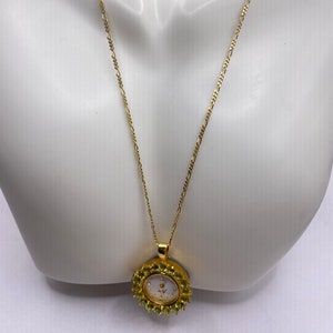 May include: A gold-toned necklace featuring a pendant watch. The watch has a white face with gold-toned hands and is surrounded by small, green gemstones. The necklace chain is delicate and also gold-toned.