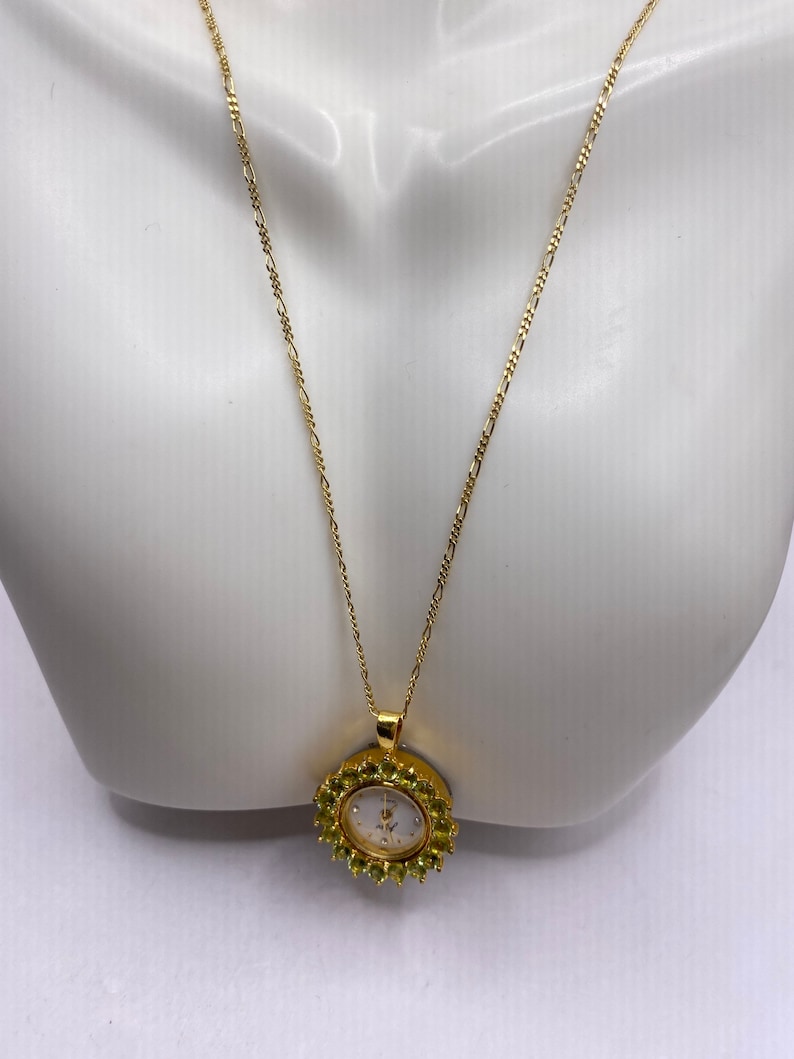 May include: A gold-toned necklace featuring a pendant watch. The watch has a white face with gold-toned hands and is surrounded by small, green gemstones. The necklace chain is delicate and also gold-toned.