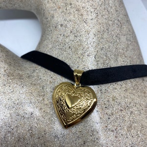 May include: A gold heart-shaped locket pendant hangs from a black velvet choker necklace. The locket has an intricate floral design.