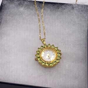 May include: A gold-toned pendant watch necklace. The watch face is white with small hour markers and hands. The watch is surrounded by green gemstones. The necklace chain is a delicate gold color. The watch is displayed in a black box with white padding.