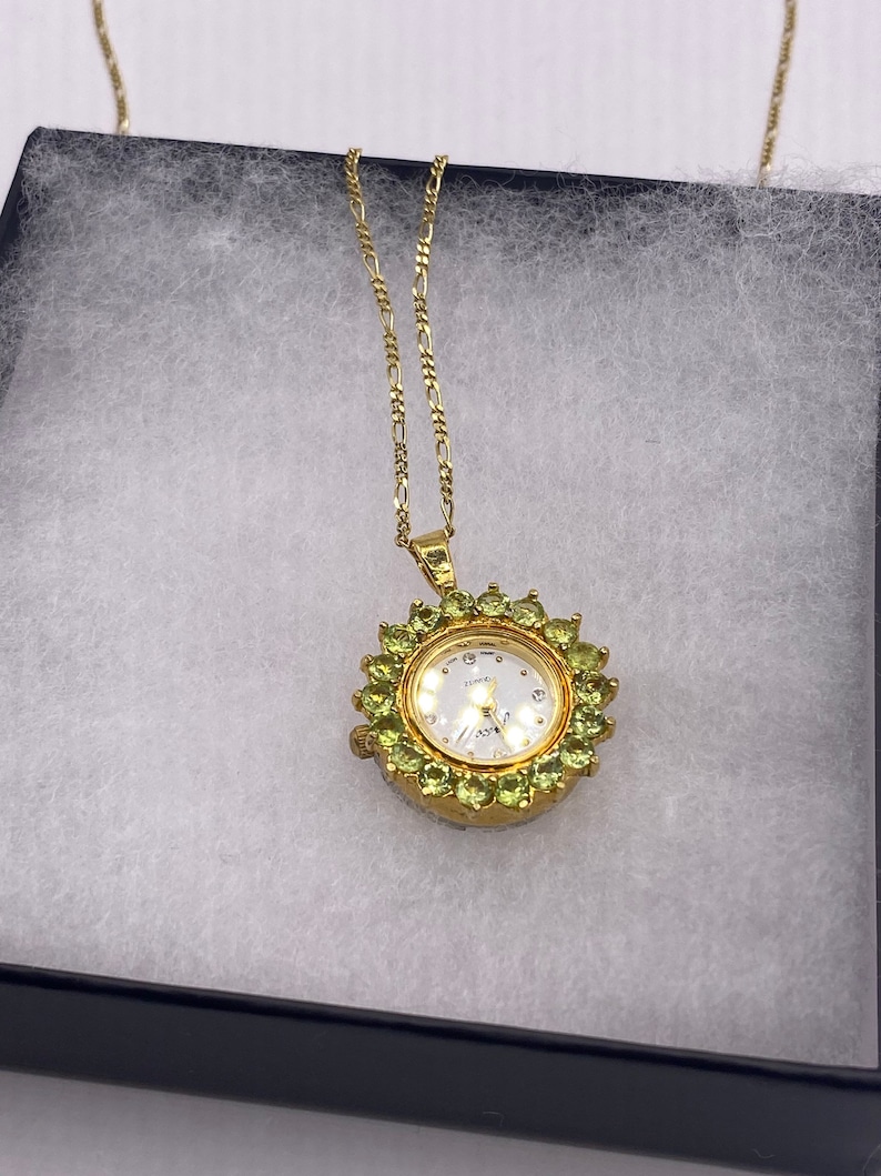 May include: A gold-toned pendant watch necklace. The watch face is white with small hour markers and hands. The watch is surrounded by green gemstones. The necklace chain is a delicate gold color. The watch is displayed in a black box with white padding.