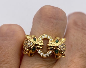 May include: A gold ring with two panther heads facing each other. The ring is set with clear and green gemstones.