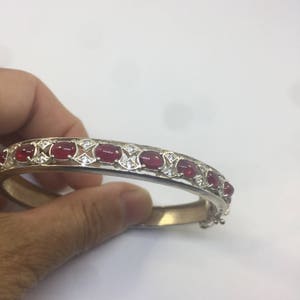 May include: A silver bangle bracelet with a row of red oval stones and small clear stones set in a delicate design.