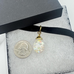 May include: A black velvet choker necklace featuring a small, teardrop-shaped pendant with an iridescent, opalescent finish and gold-toned metal hardware. A United States quarter coin is placed next to the pendant for size reference.