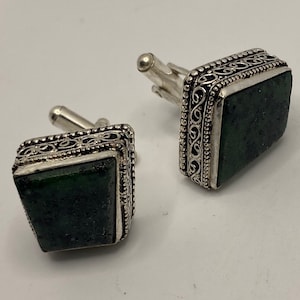 May include: A pair of silver cufflinks with green stone insets. The cufflinks have an intricate, ornate design around the edges.