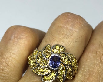 Può includere: Un anello d'argento con un design floreale. L'anello presenta una gemma blu ovale centrale, circondata da gemme gialle. I petali sono dettagliati con pietre gialle più piccole, creando un gioiello vibrante e accattivante.