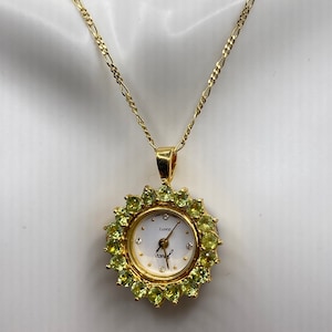 May include: Gold-toned necklace with a watch pendant. The watch face is white with small diamond accents and gold hands. The watch is surrounded by green gemstones. The chain is a delicate gold link.