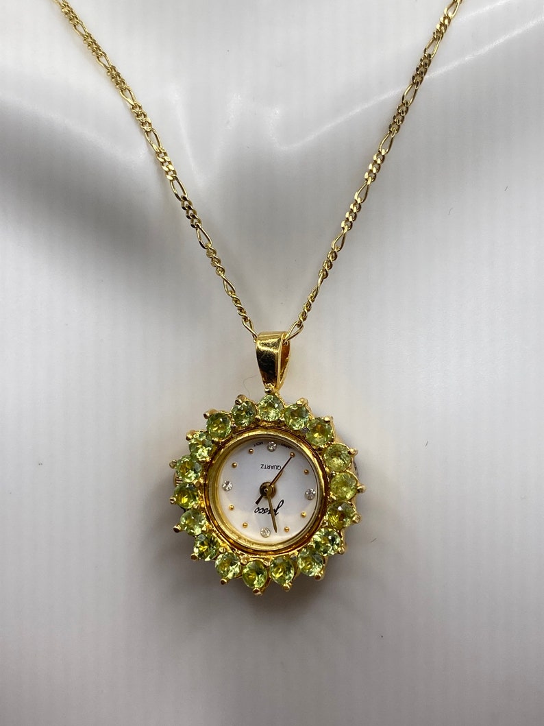 May include: Gold-toned necklace with a watch pendant. The watch face is white with small diamond accents and gold hands. The watch is surrounded by green gemstones. The chain is a delicate gold link.
