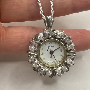 Op de afbeelding: Een zilverkleurige hangerhorlogeketting. De wijzerplaat is wit met zwarte wijzers en de naam "Jasco". Het horloge is omgeven door heldere, ovaalvormige stenen. De ketting is een zilveren touwstijl.