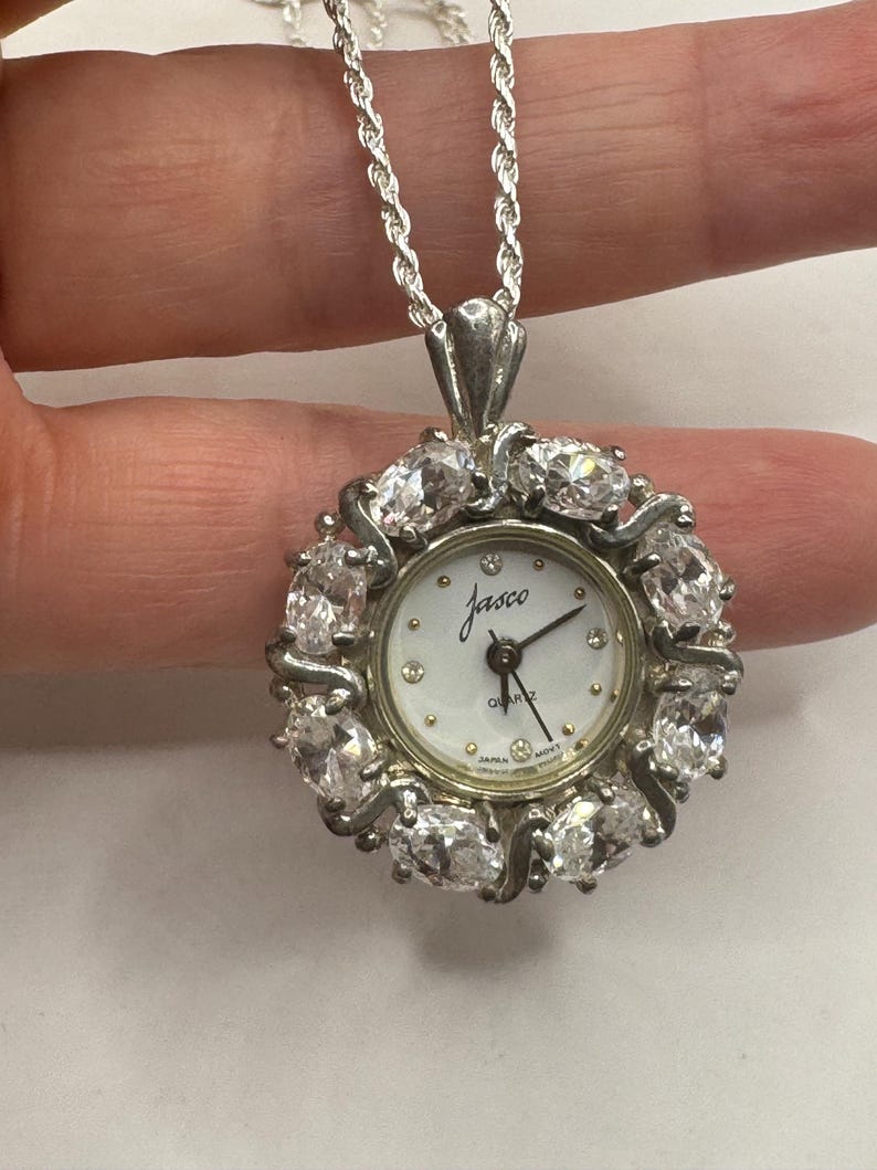 Op de afbeelding: Een zilverkleurige hangerhorlogeketting. De wijzerplaat is wit met zwarte wijzers en de naam "Jasco". Het horloge is omgeven door heldere, ovaalvormige stenen. De ketting is een zilveren touwstijl.