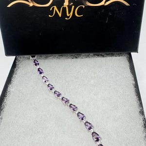 May include: A silver bracelet featuring a line of pear-shaped purple gemstones. The bracelet is presented in a black box with the inscription "Nemesis NYC" on the lid.