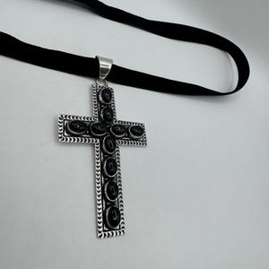 Op de afbeelding: Een zilveren kruis hanger met zwarte onyx stenen, hangend aan een zwart fluwelen lint.