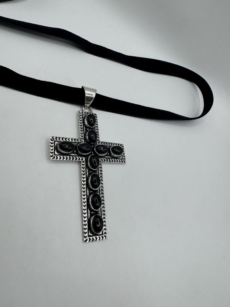Op de afbeelding: Een zilveren kruis hanger met zwarte onyx stenen, hangend aan een zwart fluwelen lint.