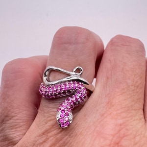 May include: A silver-toned snake ring with a wrap-around design. The snake's body is encrusted with small, bright pink gemstones. The ring is worn on a finger, showcasing the jewelry's intricate details and vibrant color.