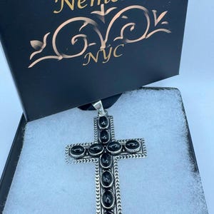 Op de afbeelding: Een zilveren kruis hanger met zwarte onyx stenen, in een zwarte doos met de tekst "www.NemesisjewelryNYC.com Nemesis NYC".