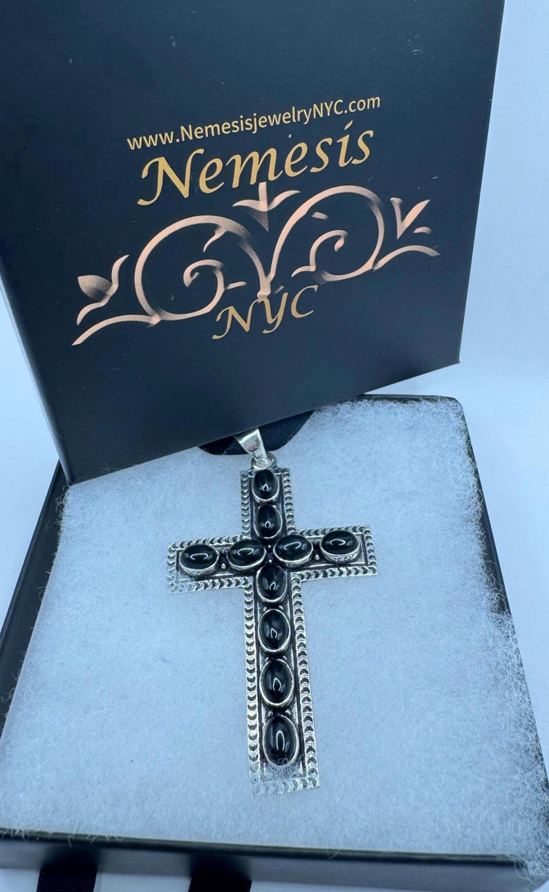 Op de afbeelding: Een zilveren kruis hanger met zwarte onyx stenen, in een zwarte doos met de tekst "www.NemesisjewelryNYC.com Nemesis NYC".