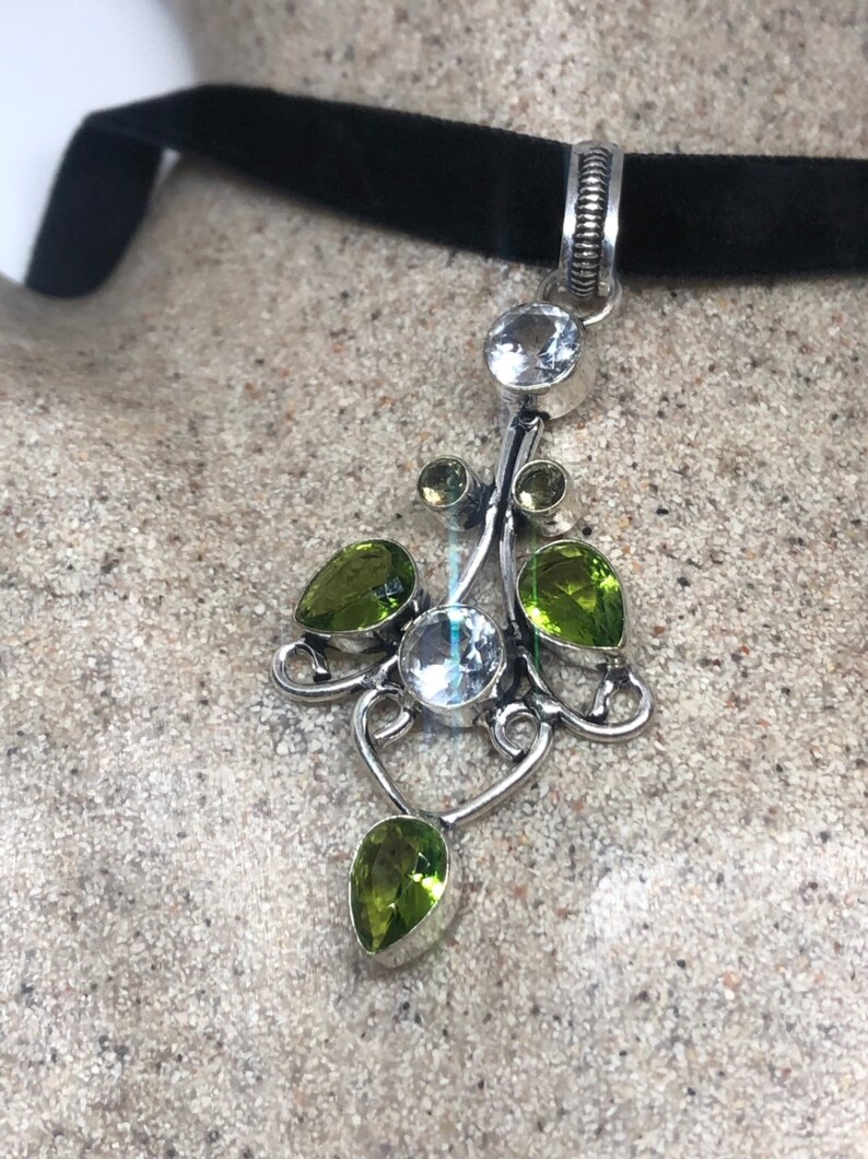 May include: A silver pendant necklace with a black velvet choker. The pendant features a delicate silver design with clear and green gemstones.