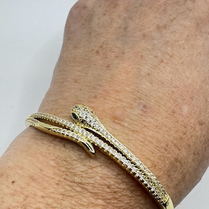 May include: Gold-toned snake bracelet with sparkling clear and green gemstones. The bracelet wraps around the wrist, featuring a snake head design. The snake's head has a small green gemstone for the eye. A stylish piece of jewelry.