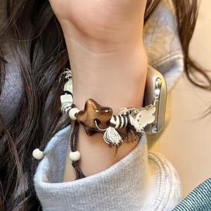 May include: A brown star-shaped charm bracelet with white and black beads, and a tassel. The bracelet is worn on a wrist next to a silver watch. The person is wearing a gray sweatshirt and blue jeans.