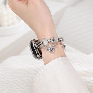 Op de afbeelding: Een zilverkleurige metalen horlogeband met diverse decoratieve bedels, waaronder een centraal hartvormig bedeltje met vleugels en heldere kristallen, bevestigd aan een zwarte smartwatch-behuizing, gedragen met een dunne zilveren ring.