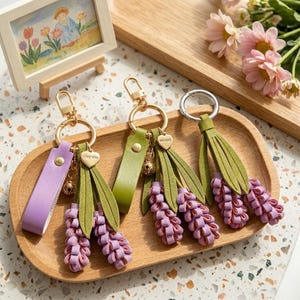 May include: Three keychains featuring lavender-colored flower clusters made of folded material, green leaves, and gold-toned metal rings. One keychain has a purple strap, another a green strap, and the third has a silver ring. Small heart charms are attached.