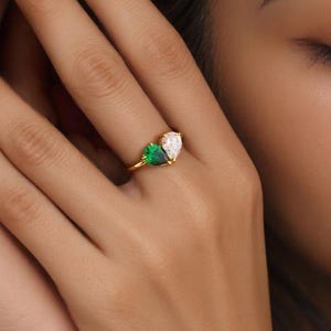 Op de afbeelding: Een gouden ring met een peervormige smaragd en een peervormige diamant. De ring heeft een delicate gouden band. De edelstenen worden prominent weergegeven, wat een opvallend contrast creëert.