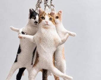 Peut inclure: Un ensemble de trois ornements en tissu en forme de chat, chacun présentant un pelage distinct incluant noir et blanc, tabby brun et blanc, et orange et blanc, tous suspendus par des cordons noirs avec des fermoirs en métal argenté.