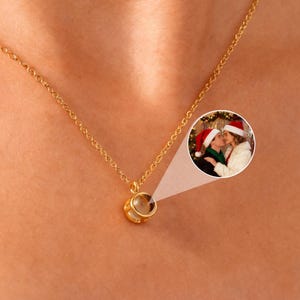 May include: A gold-coloured necklace featuring a small, round pendant. The pendant holds a small, circular image of two people wearing Santa hats. The necklace is worn around a person's neck.