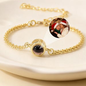 May include: A gold-colored bracelet with a round, dark-colored center stone. The bracelet is on a white plate. A small inset image shows two people wearing Santa hats. The bracelet has a delicate chain design.