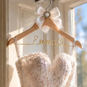 Peut inclure: Une robe de mariée bustier en dentelle blanche avec un décolleté en cœur suspendue à un cintre en bois. Le cintre présente un nœud en satin blanc avec une perle centrale et une inscription en fil doré indiquant Emma avec un petit cœur.