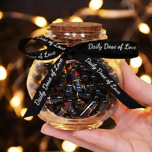 Op de afbeelding: Een heldere glazen pot gevuld met zwarte capsules, sommige met rode hartaccenten, en een kurken deksel. Een zwart lint met de tekst "Daily Dose of Love" is om de pot gebonden. De achtergrond heeft vage, warmgekleurde lichten.
