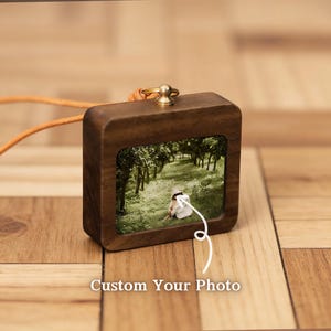 May include: A wooden pendant with a small photo inside, featuring a person in a white hat sitting in a grassy area with trees. The pendant has a gold-colored metal loop and an orange leather cord. The text "Custom Your Photo" is at the bottom.
