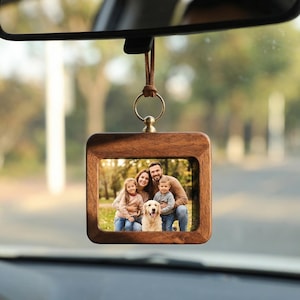 May include: A wooden car charm with a family photo inside. The charm is square with rounded corners and hangs from a leather cord. The photo shows a family of four with a dog, all smiling.
