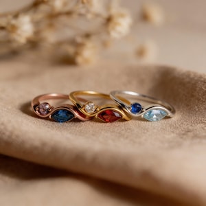 Könnte beinhalten: Vier Edelsteinringe in Roségold, Gold und Silber. Jeder Ring hat eine einzigartige Kombination aus Edelsteinen, darunter rosa, blaue, rote und klare Steine. Die Ringe werden auf einem neutralen Stoff präsentiert, der ihr elegantes Design hervorhebt.