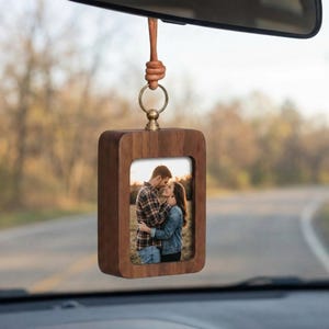 May include: A wooden car charm with a rectangular frame holding a photograph of a couple. The charm is suspended by a leather cord and a gold-colored ring. The frame is made of dark wood.