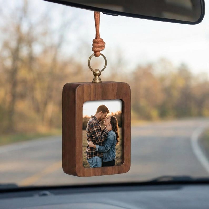 May include: A wooden car charm with a rectangular frame holding a photograph of a couple. The charm is suspended by a leather cord and a gold-colored ring. The frame is made of dark wood.