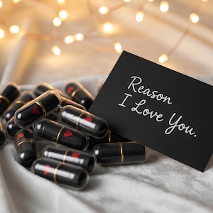 Könnte beinhalten: Eine Ansammlung schwarzer Kapseln mit goldenen Akzenten und einem roten Herzdesign im Inneren, die auf einer weißen Oberfläche liegen. Eine schwarze Karte mit dem Text "Reason I Love You" ist in der Nähe aufgestellt. Weiche, warme Lichter im Hintergrund.