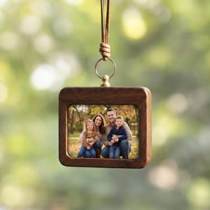 Könnte beinhalten: Ein quadratischer Holzfotorahmen mit einem Familienporträt im Inneren, der an einer braunen Lederschnur hängt. Der Rahmen hat eine dunkelbraune Oberfläche und einen goldfarbenen Metallring oben. Der Hintergrund ist verschwommen grün.