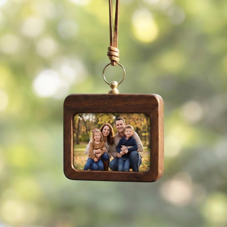 May include: A square, wooden photo frame with a family portrait inside, suspended by a leather cord. The frame has a dark brown finish and a gold-colored metal ring at the top. The background is a blurred green and yellow.