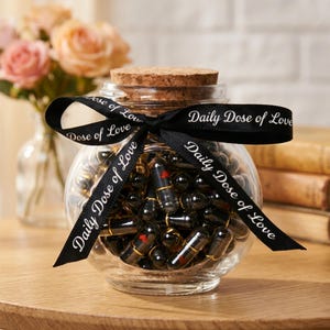 Könnte beinhalten: Ein klares Glasgefäß gefüllt mit schwarzen Kapseln, gebunden mit einem schwarzen Band mit weißem Aufdruck "Daily Dose of Love". Das Glas hat einen Korkdeckel. Blumen im Hintergrund.