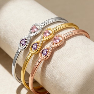 Könnte beinhalten: Drei Armbänder mit Unendlichkeitszeichen in Silber, Gold und Roségold. Jedes Armband hat einen herzförmigen rosa und lila Edelstein. Die Namen "Smith" und "Johnson" sind eingraviert. Präsentiert auf hellem Stoff.